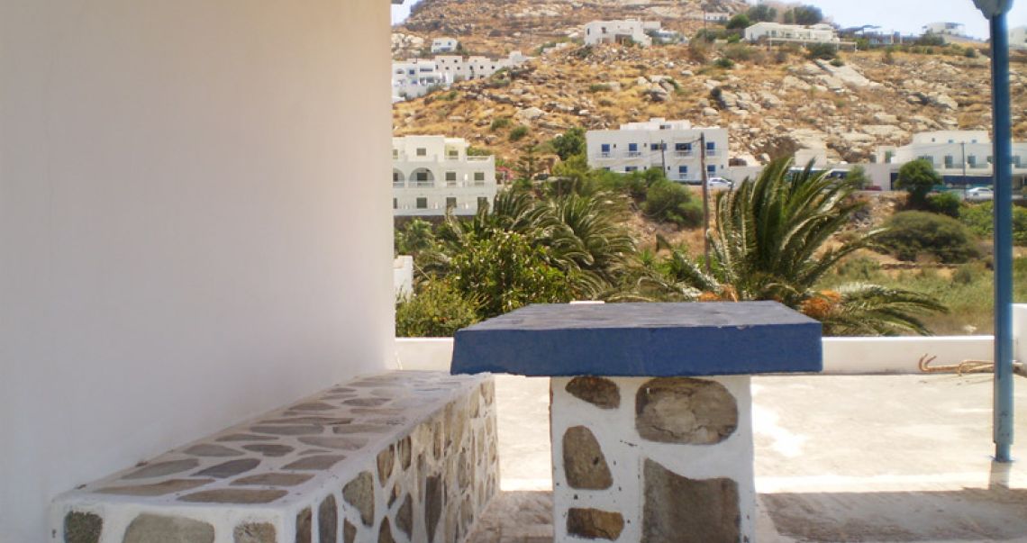 Shaded outdoor terrace with a stone table and bench under a wooden pergola, overlooking the Mediterranean landscape.