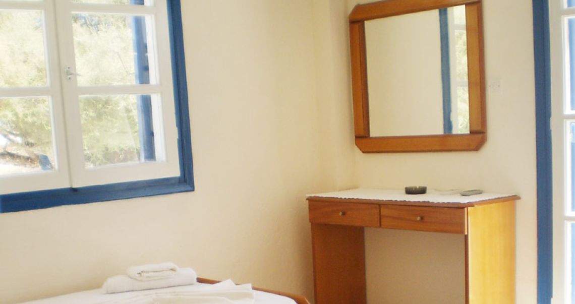 Hotel room interior showing a wooden vanity desk with a mirror, located next to a bright window with blue frames.