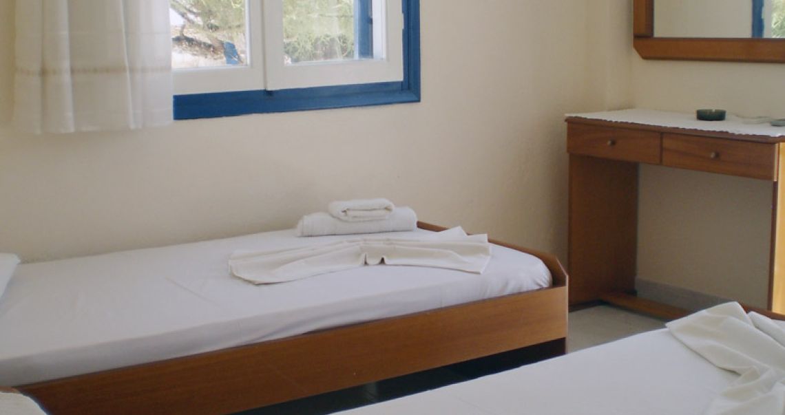 Traditional twin hotel room in Ios featuring wooden furniture, fresh white linens, and a classic blue window frame.
