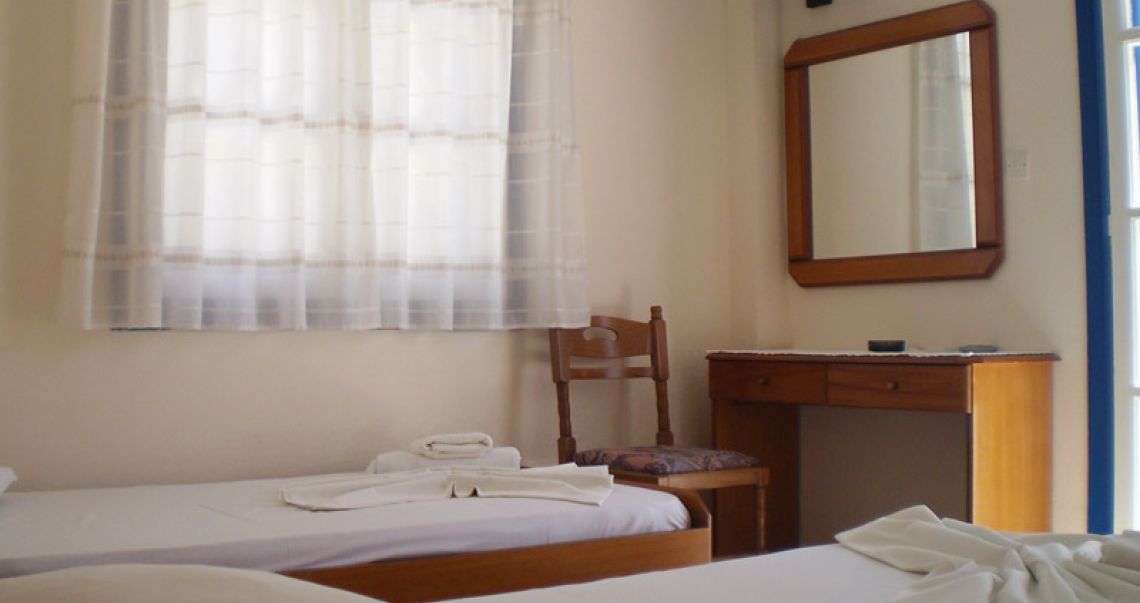 Bright hotel room interior with twin beds, a wooden vanity desk, wall-mounted TV, and light-filtering curtains.
