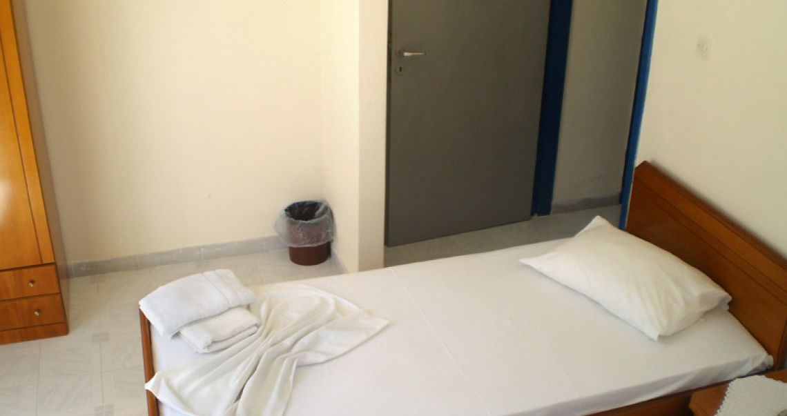 Simple and clean guest room featuring a single wooden bed, a large wardrobe, and cool tiled flooring.