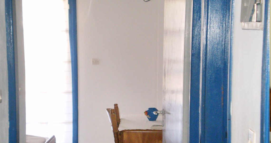 View through a traditional blue door frame into a bright hotel room with a workspace and sunlit balcony.