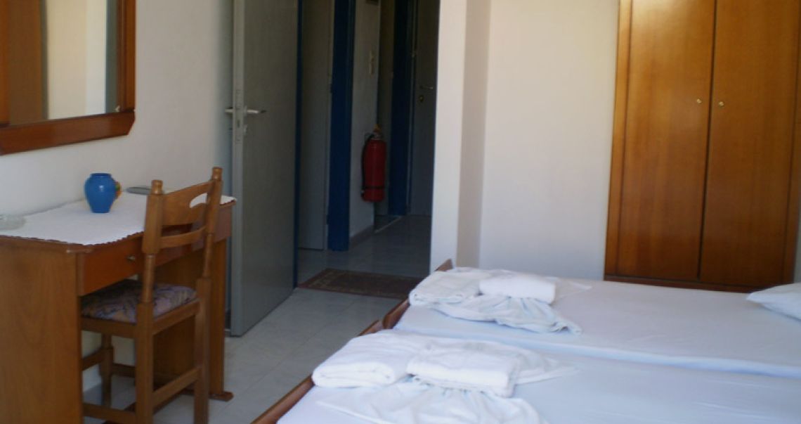 Spacious hotel room layout showing twin beds, a wooden wardrobe, a vanity mirror, and the entrance hallway.