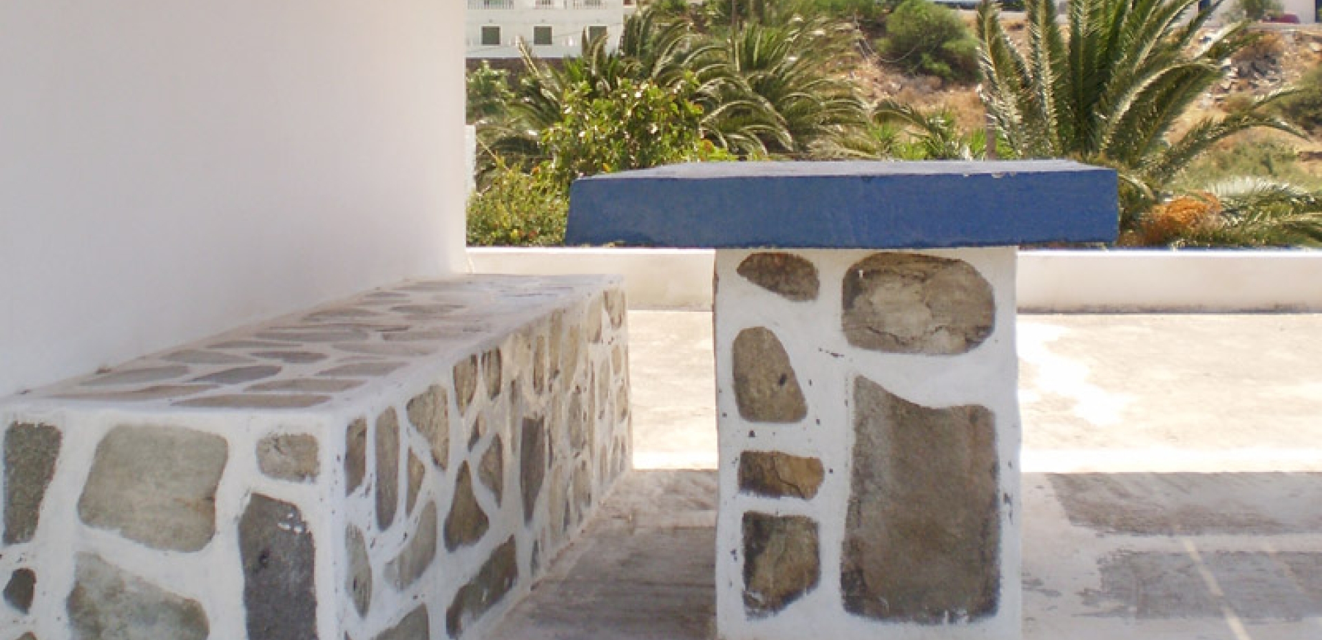 Outdoor seating area featuring a traditional stone-built bench and a blue-topped table with a view of palm trees and hills.